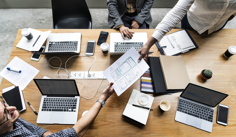 Von oben fotografiert: Ein Schreibtisch, an dem drei Personen an den Laptops arbeiten. Eine Person reicht der anderen gerade ein Dokument über.