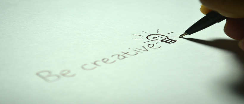 Auf dem Bild ist auf einem türkisen Papier das Wort creative drauf geschrieben mit einer Glühbirne daneben gezeichnet. Jemand hält einen Stift mit der Hand und schreibt gerade diese Worte.